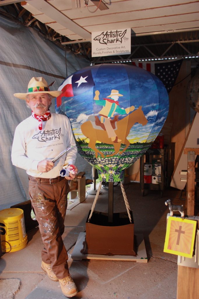 Artist in hat next to fabricated hot air balloon with painting of man riding a horse.